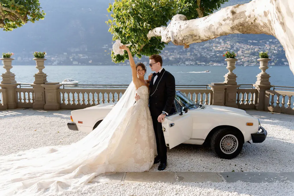 A newly married couple beside a vintage car overlooking Lake Como